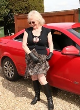 In these porn photos, a naked old lady with glasses poses next to a red car and caresses her body